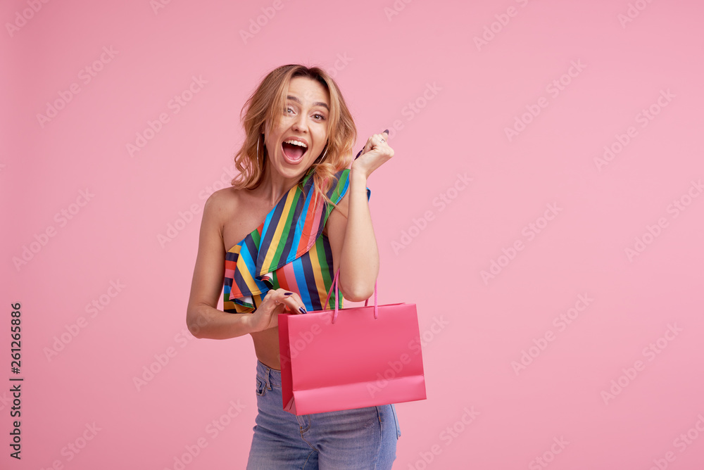 Fototapeta premium Portrait of an excited beautiful girl wearing colorful clothes holding shopping bag isolated over pink background. Place to sign or text on the package