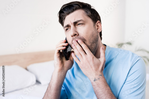 handsome man yawning while talking on smartphone in bedroom