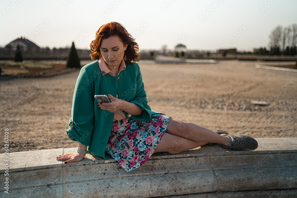 Young woman uses phone in a palace park sitting on a fountain