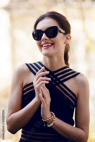 Fashionable look at cheerful woman with sublime from, wearing in a elegant black dress and sunglass, in daytime posing on the balcony.