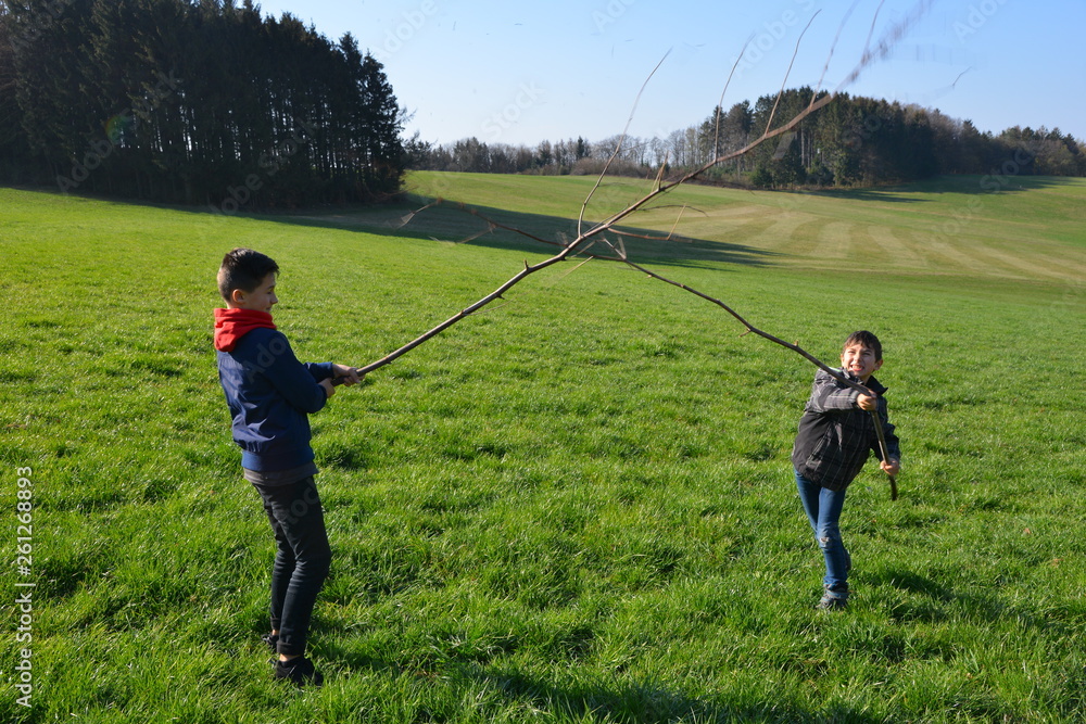 Outdoor-Abenteuer mit Kindern: Jungs beim Stockkampf mit Ästen auf der grünen Wiese im ländlichen Odenwald, Hessen, Deutschland