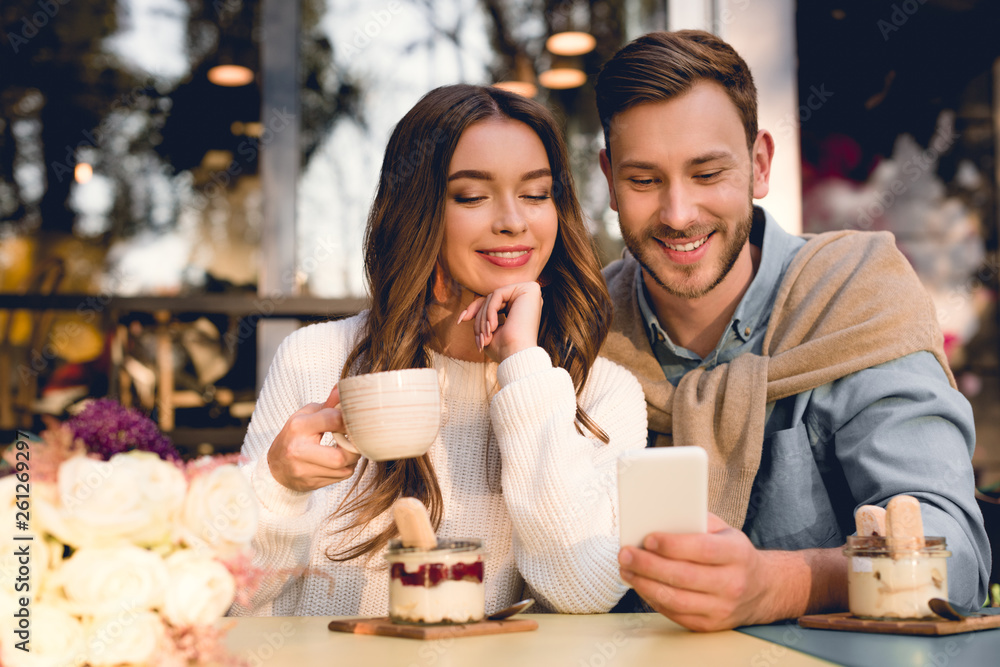 cheerful man looking at smartphone near attractive girl holding cup of coffee cheerful man looking at smartphone near attractive girl holding cup of coffee