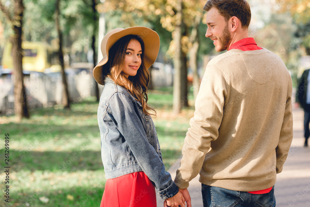 Fototapeta premium handsome man looking at attractive girlfriend in hat while holding hands in park