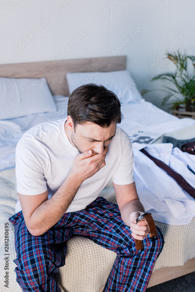 yawning man holding watch while sitting on bedding at home yawning man holding watch while sitting on bedding at home