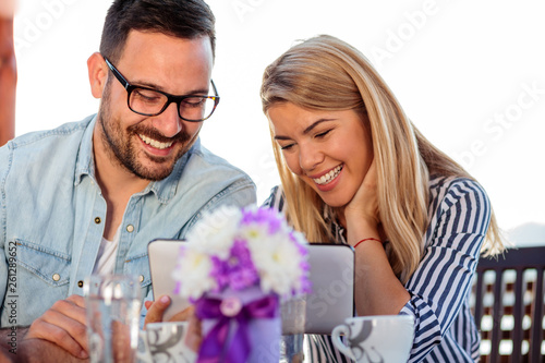 Smiling happy young couple browsing internet or looking at pictures on a tablet, drinking coffee in an outdoor cafe