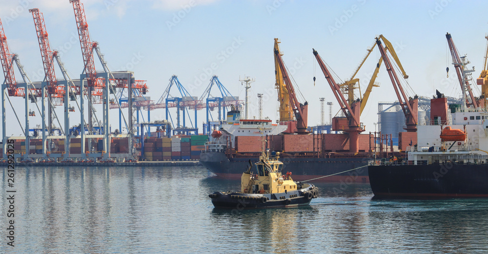 Tugboat at the bow of cargo ship , assisting the vessel to maneuver in Sea Port of Odessa. Lifting cargo cranes, ships and grain dryer in  Port