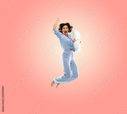 fun, people and bedtime concept - happy young woman full of energy in blue pajama holding pillow and jumping over living coral pink background