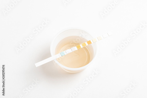 A set of urine sample on a round plastic cup with a single unused urine reagent strip against a plain white background.