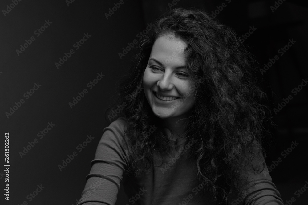 Black and white authentic horizontal portrait of cheerful Caucasian curly haired female model, smiling, looking aside,having conversation with friend, posing in dark room indoors. Beauty concept. Black and white authentic horizontal portrait of cheerful Caucasian curly haired female model, smiling, looking aside,having conversation with friend, posing in dark room indoors. Beauty concept.