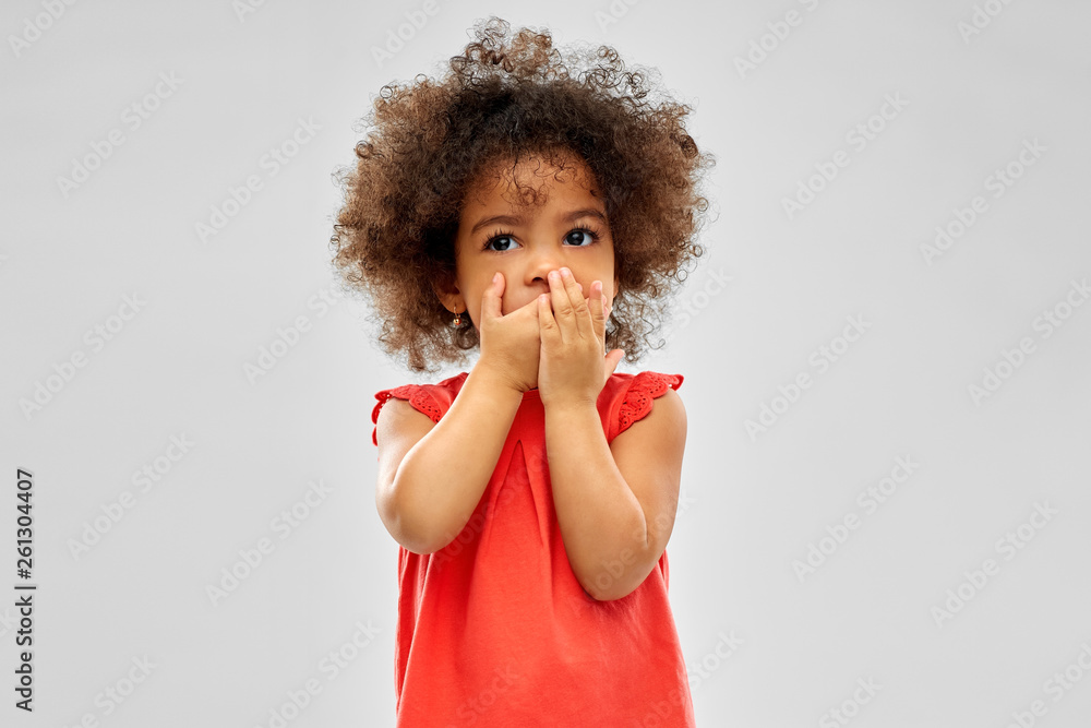 childhood, expression and emotion concept - confused little african american girl covering mouth by hands over grey background - obrazy, fototapety, plakaty 