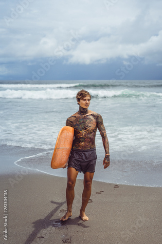 Photography Attractive male surfer in tattoos on the ocean with an orange bright longboard surfboard