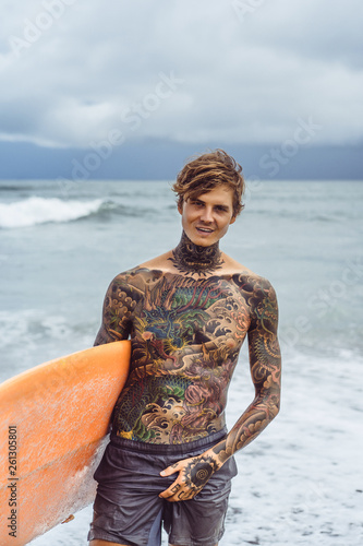 Photography Attractive male surfer in tattoos on the ocean with an orange bright longboard surfboard