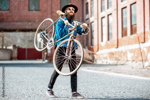 Fototapeta Naklejka Na Ścianę i Meble -  Lifestyle portrait of a bearded hipster dressed stylishly with hat and jacket carrying his retro bicycle on the urban background
