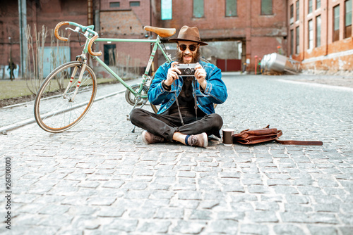 Bearded hipster dressed stylishly with hat and jacket sitting with photo camera and retro bicycle on the street outdoors, wide view