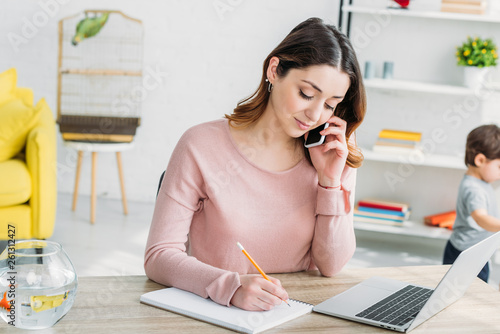 beautiful woman talking on smartphone while sitting at table with laptop at home photo