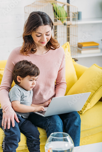 attractive mother with cute son using laptop while sitting on yellow sofa at home