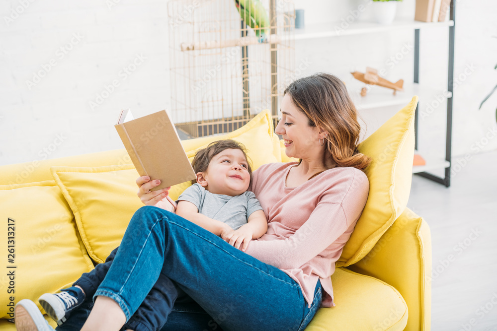 cheerful mother with book and cute smiling boy resting on yellow sofa at home