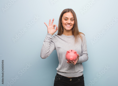 Young pretty caucasian woman cheerful and confident doing ok gesture. She is holding a piggy bank.