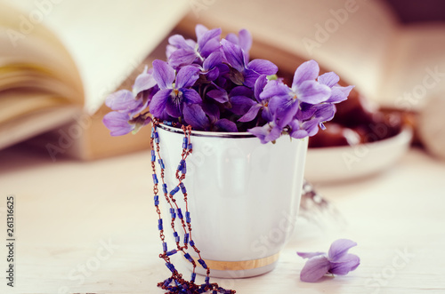 Still life with violet in white cup  old books and date fruits in a plate. Romantic floral background.