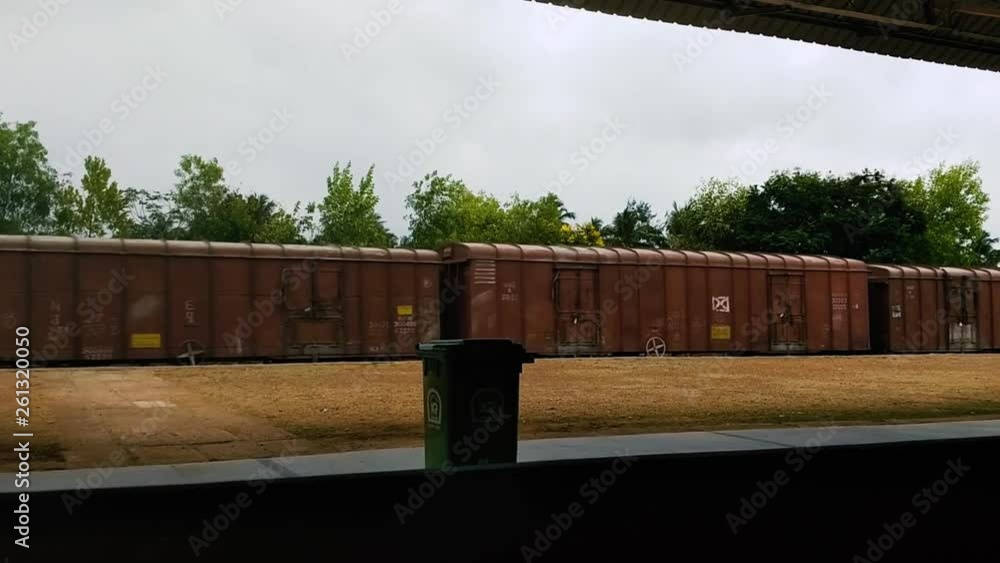 Brown cars of a long freight train speeding down the tracks past a railroad station in India. Green recycling bin in frame as a good train races past. Brown cars of a long freight train speeding down the tracks past a railroad station in India. Green recycling bin in frame as a good train races past.