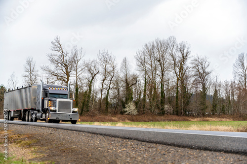 Black big rig classic semi truck with turned on headlight transporting commercial cargo in bulk semi trailer running on the road