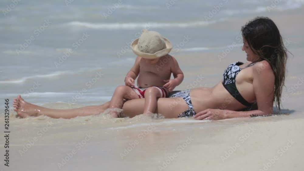 Mom and toddler boy, playing together on the beach edge, water splashing them Mom and toddler boy, playing together on the beach edge, water splashing them