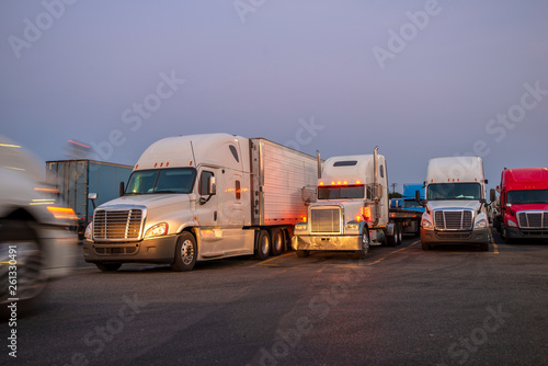 Different big rigs semi trucks with semi trailers standing in row on the twilight truck stop and illuminated by the light of passing truck