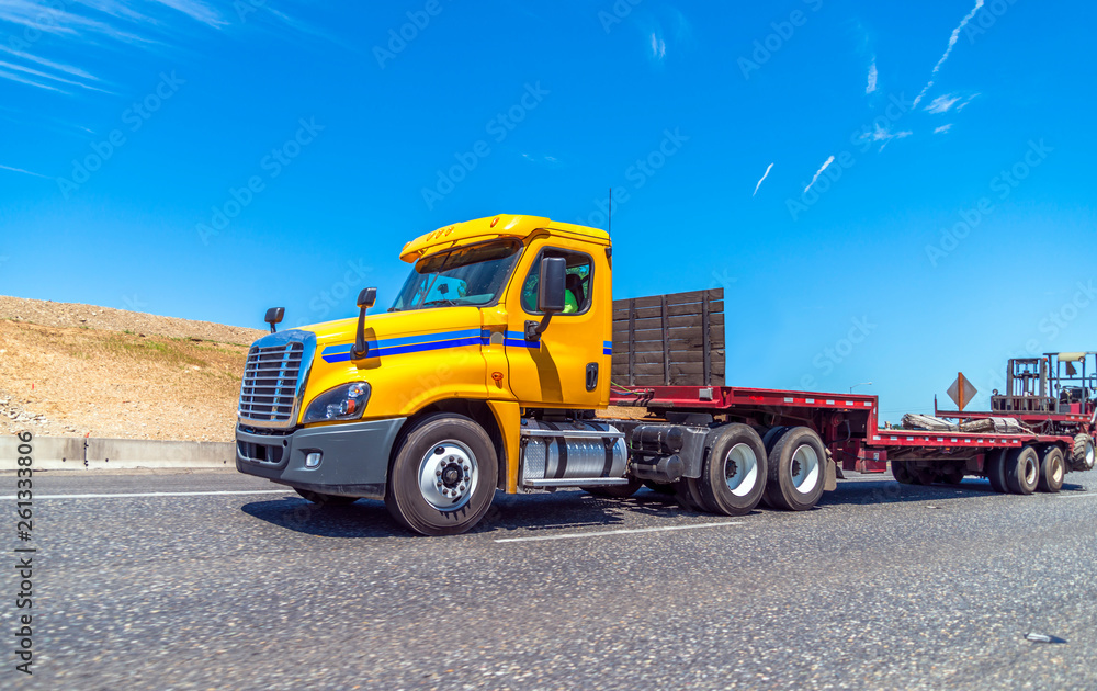 Yellow big rig day cab semi truck with step down semi trailer and forklift mounted at the end of the trailer running on the road to warehouse for loading