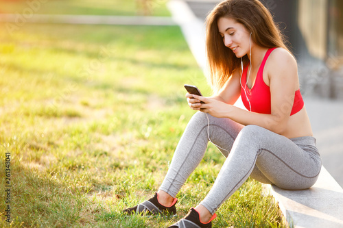 Young sporty woman resting in the park after training, listening to music through headphonesand using the phone. © shootsroom