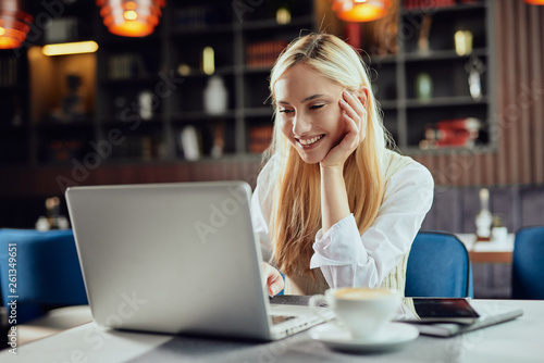Beautiful smiling Caucasian blonde female blogger dressed smart casual sitting in cafeteria and using laptop.