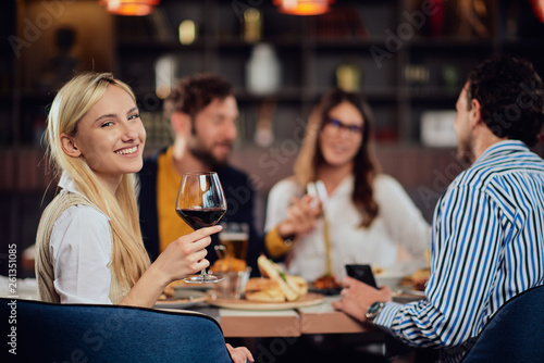 Portrait of beautiful Caucasian blonede woman sitting in restaurant and holding glass of red wine. In background friends having dinner.