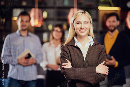 Portrait of beautiful Caucasian blonde businesswoman standing at restaurant with arms crossed. In background her colleagues standing and posing.