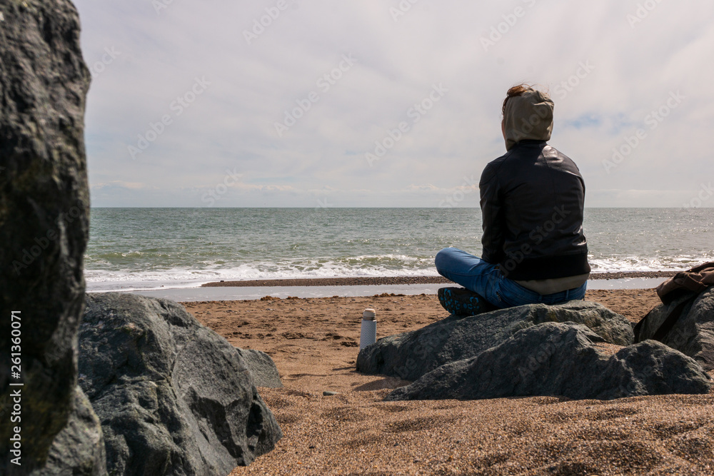 Young woman relaxing and meditating at the seashore on a rocky beach. Moody winter day seascape in Greystones, Wicklow, Ireland. Young woman relaxing and meditating at the seashore on a rocky beach. Moody winter day seascape in Greystones, Wicklow, Ireland.
