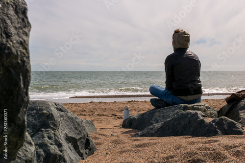 Young woman relaxing and meditating at the seashore on a rocky beach. Moody winter day seascape in Greystones, Wicklow, Ireland.