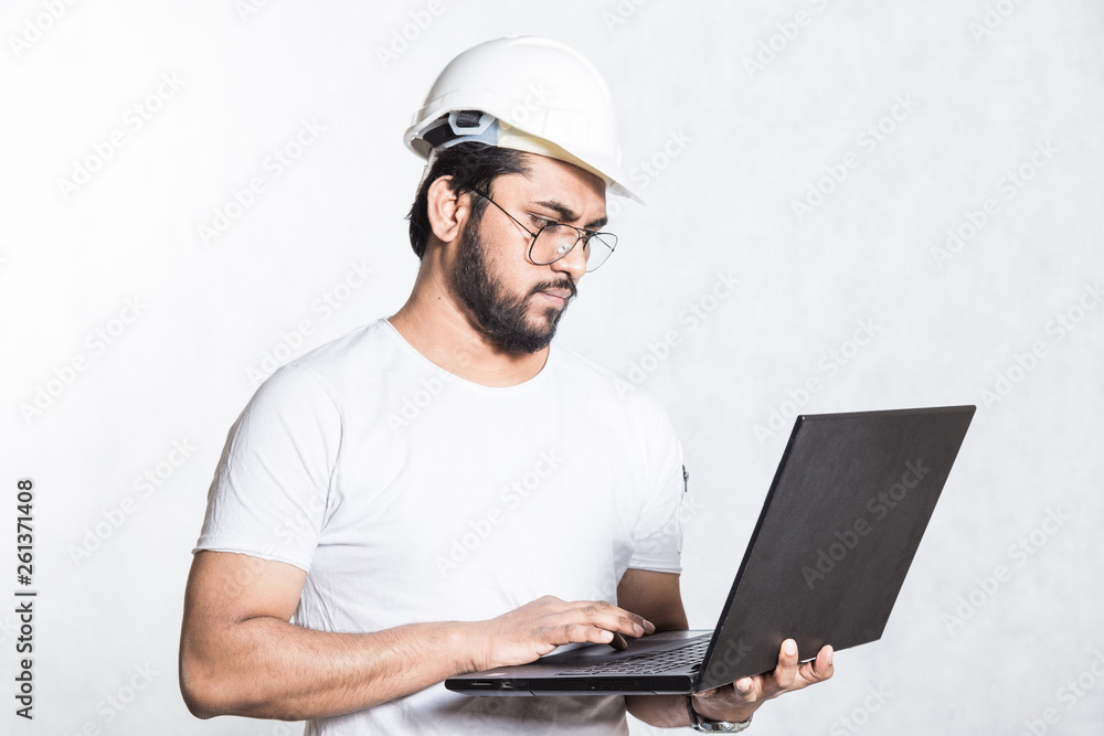 Young builder engineer in glasses and a white safety helmet, stands with an open laptop.