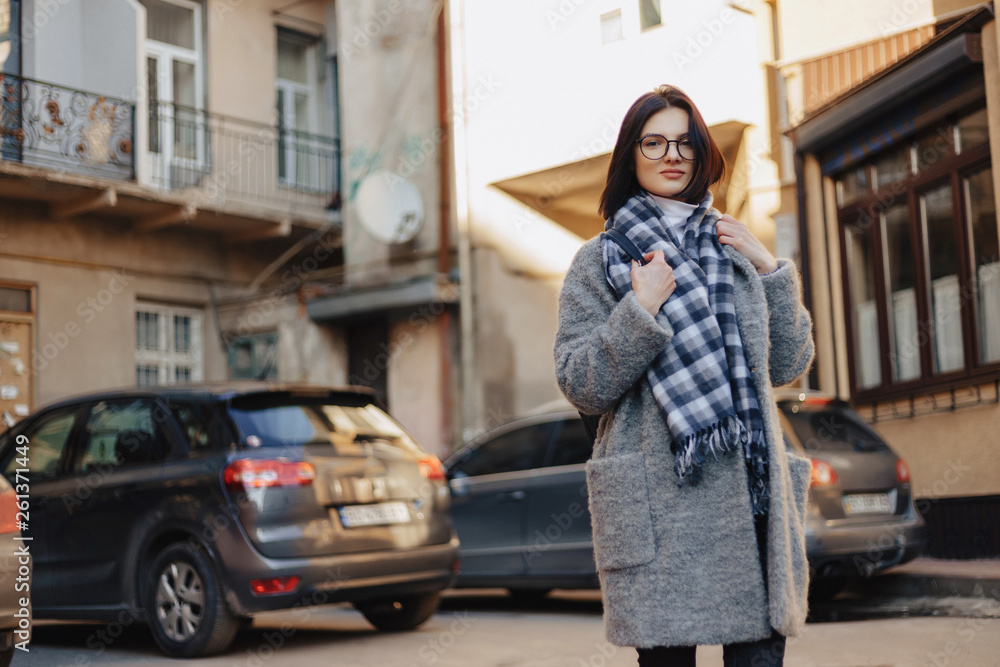 Attractive positive young girl wearing glasses in a coat on the background of buildings on cars