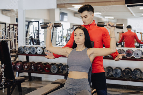 A young girl wearing sportswear in a gym performs dumbbell exercises, the coach helps her