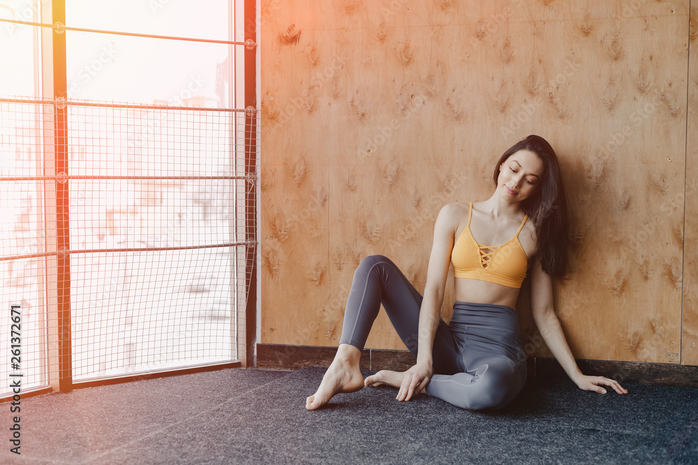 Young attractive fitness girl sitting on the floor near the window on the background of a wooden wall, resting on yoga classes Young attractive fitness girl sitting on the floor near the window on the background of a wooden wall, resting on yoga classes