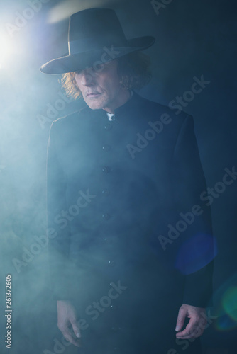 Mysterious victorian priest in black coat and hat in fog.