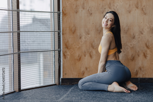 Young attractive fitness girl sitting on the floor near the window on the background of a wooden wall, resting on yoga classes