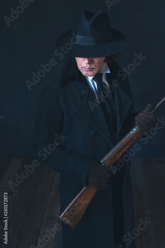 Victorian man in long black coat and hat holding rifle.