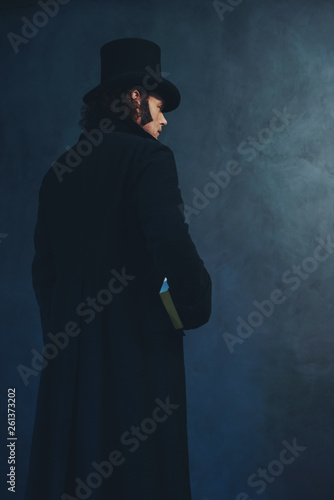 Edwardian man in long black coat and hat holding book standing towards grey wall. photo