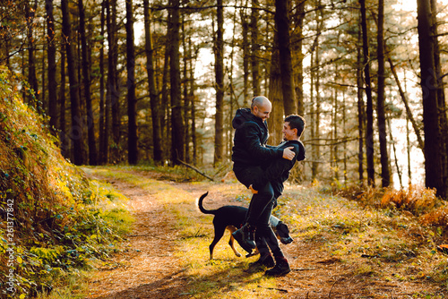 Side view of homosexual couple embracing and kissing near dog on route in forest in sunny day photo