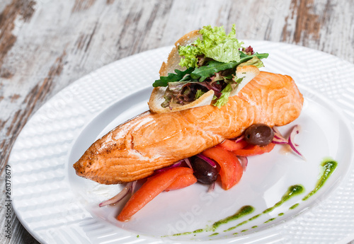 Baked salmon garnished with olives, greens, tomatoes on plate over wooden background. Hot fish dish