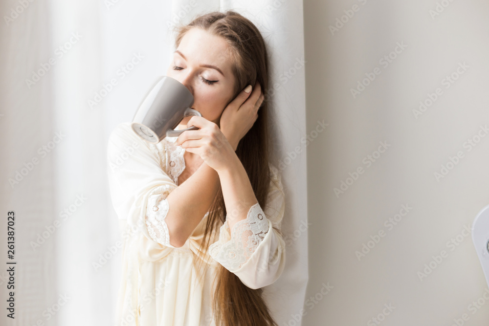 Attractive young girl in nightwear stands at the window and drinks coffee. Morning mood Attractive young girl in nightwear stands at the window and drinks coffee. Morning mood