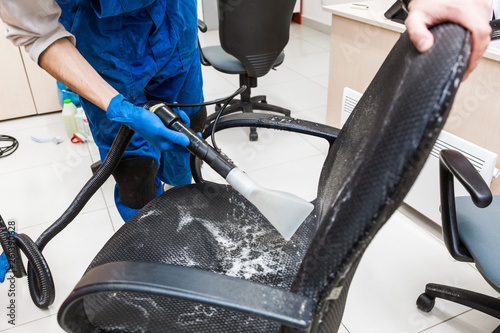 Young man in workwear and rubber gloves cleans the office chair with professional equipment.