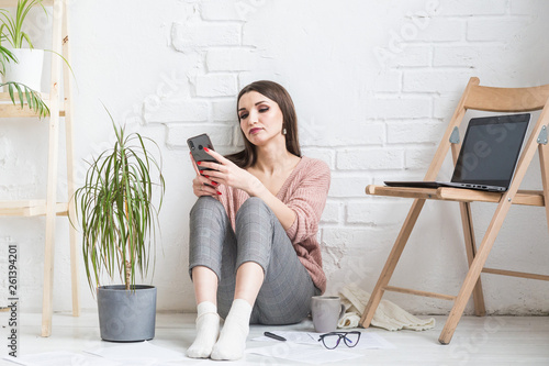A happy woman in homemade casual clothes sits in a bright interior and uses a telephone, the girl is resting while working at a laptop and surfing the Internet. Cozy freelance work space,