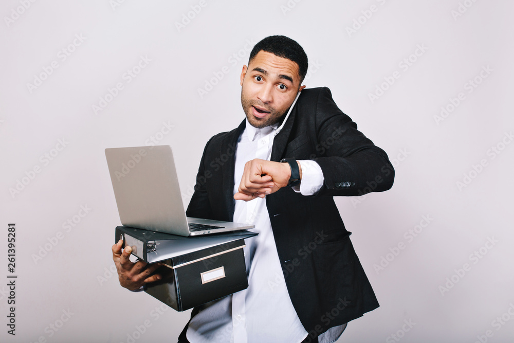 Busy hard-working guy in white shirt and black jacket talking on phone, holding folders, laptop, looks astonished at watch on white background. Office worker, modern technology, career Busy hard-working guy in white shirt and black jacket talking on phone, holding folders, laptop, looks astonished at watch on white background. Office worker, modern technology, career