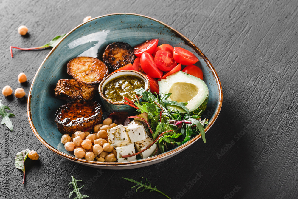 Healthy Buddha bowl dish with avocado, tomato, cheese, chickpea, fresh arugula salad, baked potatoes and sauce pesto in black background. Dieting food, clean eating Healthy Buddha bowl dish with avocado, tomato, cheese, chickpea, fresh arugula salad, baked potatoes and sauce pesto in black background. Dieting food, clean eating