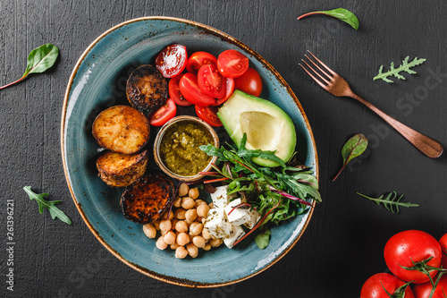Healthy Buddha bowl dish with avocado, tomato, cheese, chickpea, fresh arugula salad, baked potatoes and sauce pesto in black background. Dieting food, clean eating, top view, flat lay, copy space